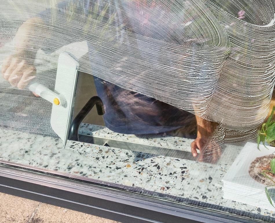 Osoba używa myjki do okien, czyszcząc szybę z widocznymi smugami. W tle widać fragment parapetu z rośliną.