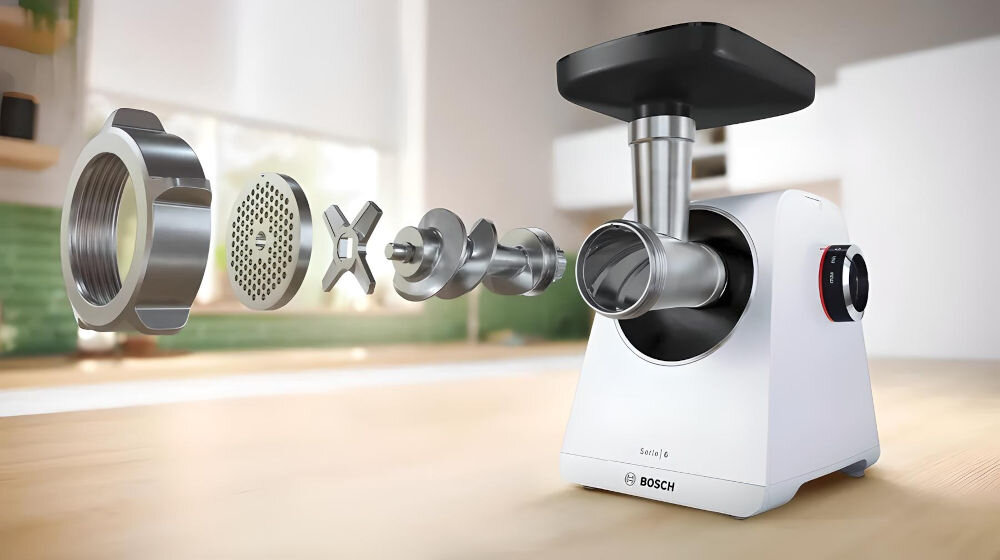 Na obrazie widoczna jest biała maszynka do mięsa z logo BOSCH, z rozłożonymi elementami metalowymi, takimi jak sitko i noże. W tle znajduje się rozmyte wnętrze kuchni.