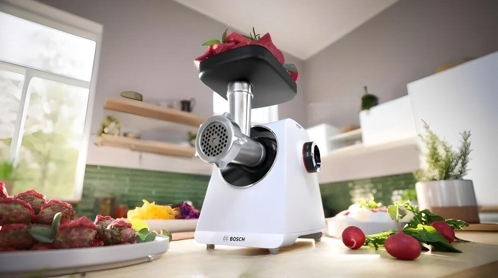 Na blacie kuchennym stoi biała maszynka do mięsa z logo BOSCH, z której wychodzi zmielone mięso. W tle widać kuchnię z zielonymi kafelkami i oknem.