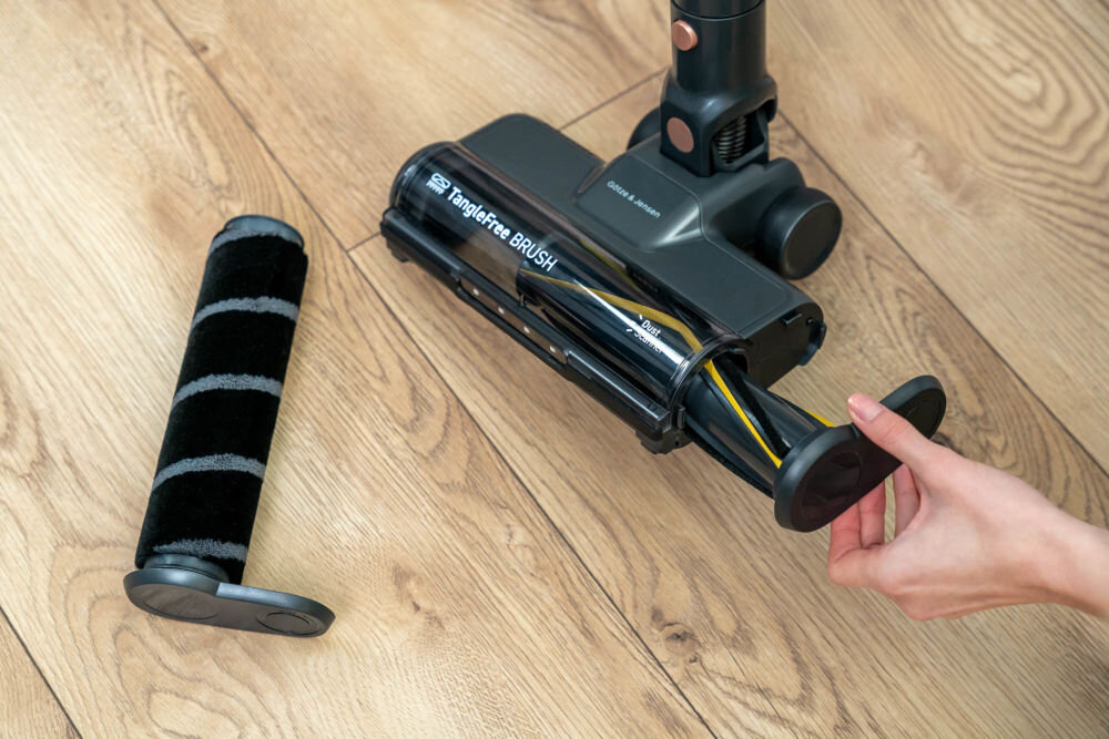 Na drewnianej podłodze znajduje się szczotka odkurzacza z napisem \'Tanglefree BRUSH\'. Obok leży wymienny wałek, a ręka trzyma element szczotki.