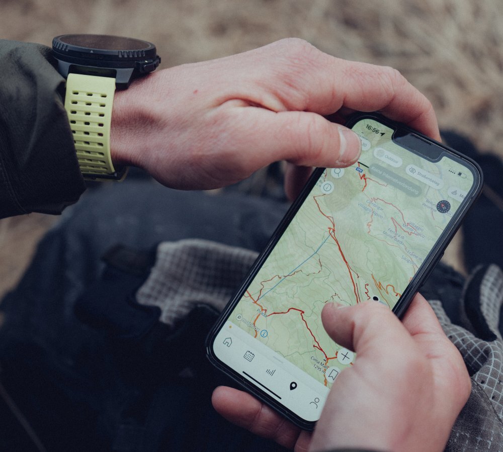 Osoba trzyma smartfon z wyświetloną mapą topograficzną, a na nadgarstku ma zegarek z jasnym paskiem. Na ekranie telefonu widoczne są szczegóły mapy oraz ikony nawigacyjne.