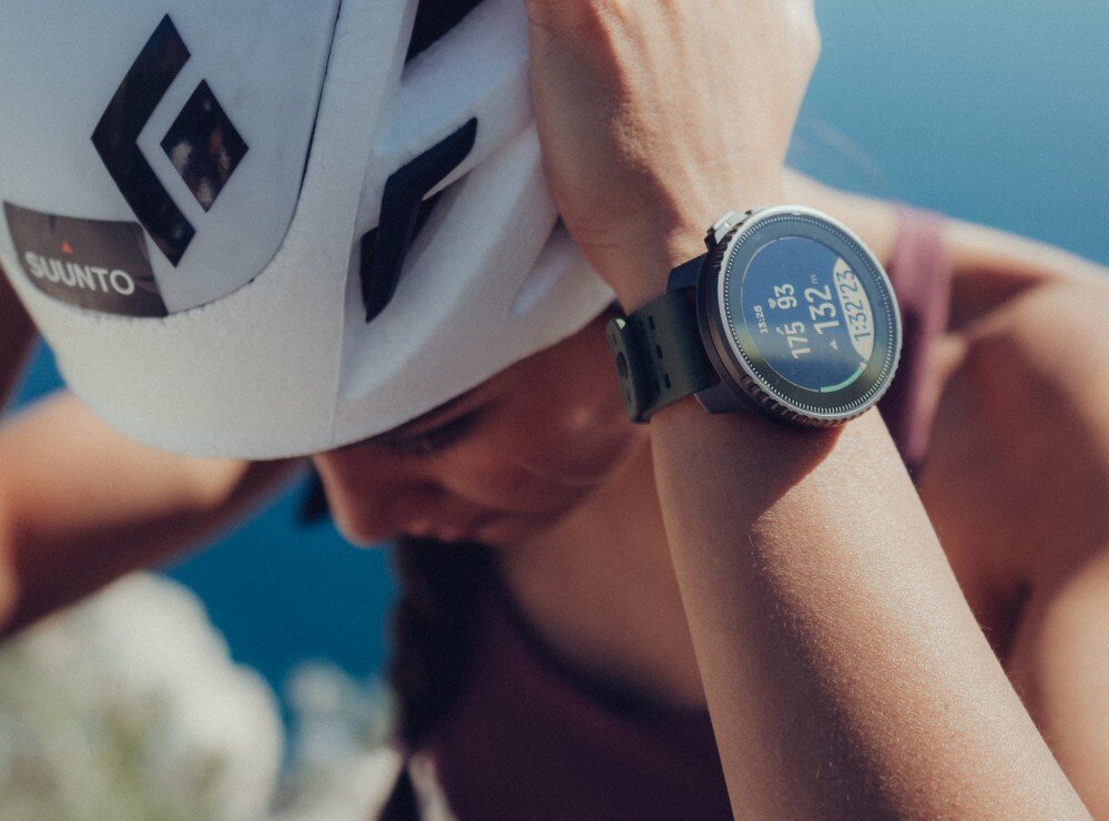 Osoba nosi biały kask z logo \'SUUNTO\' oraz smartwatch na nadgarstku z wyświetlaczem pokazującym dane cyfrowe.
