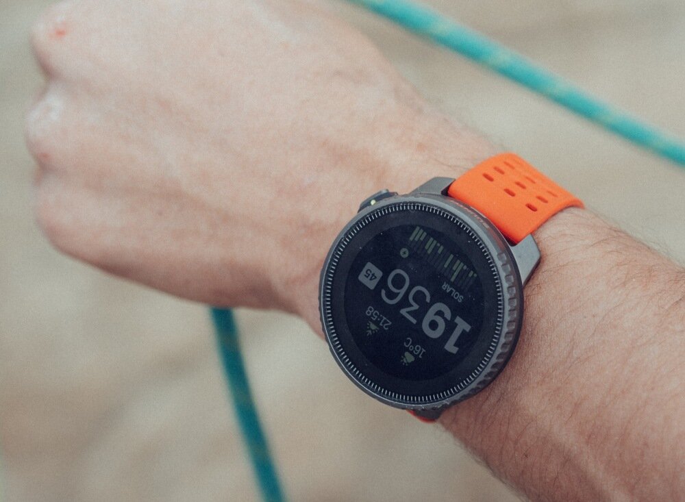 Na zdjęciu widoczny jest smartwatch z pomarańczowym paskiem na nadgarstku. Na ekranie zegarka wyświetla się liczba 1936 oraz inne dane w formie cyfrowej.