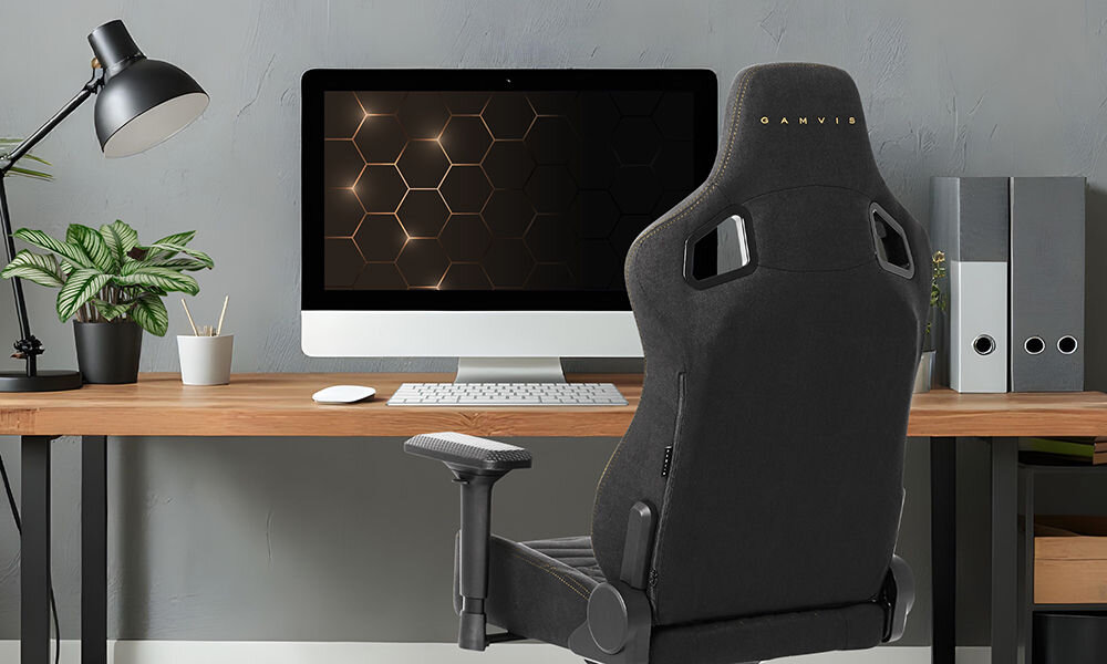 Fotel GAMVIS Elite 2.0 Pro XL Tkanina Czarny (złote elementy) prezentacja fotela w pokoju przy biurku na nim monitor i akcesoria komputerowe kwiatek lampka brązowy blat biurka detale płaskie siedzisko ZeroStress+ dekoracyjne przeszycia welurowe poduszki wsparcie wygodnej pozycji