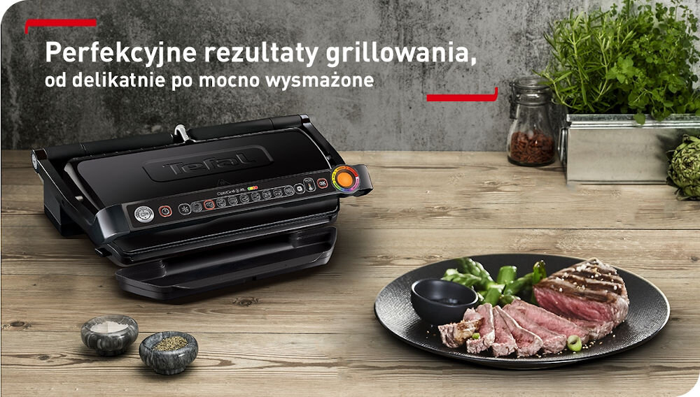 Na drewnianym blacie stoi czarny grill elektryczny z widocznym logo Tefal i kolorowymi przyciskami. Obok znajduje się talerz z grillowanym mięsem i warzywami oraz słoiki z przyprawami i ziołami, a w tle widoczny jest napis: \'Perfekcyjne rezultaty grillowania, od delikatnie po mocno wysmażone\'.
