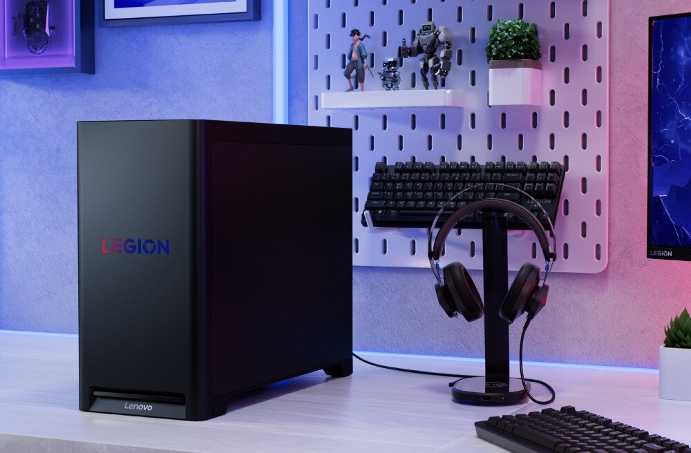 Komputer LENOVO Legion T5 30IAX10 Czarny komputer stacjonarny z logo 'LEGION' na przodzie stoi na jasnym biurku obok stojaka z zawieszonymi słuchawkami, klawiatury i monitora, opis