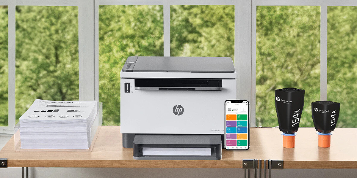 Na drewnianym stole znajduje się urządzenie wielofunkcyjne z logo HP, obok którego leży smartfon z kolorowym ekranem oraz dwa pojemniki z tuszem oznaczone jako \'15X\' i \'15A\'. W tle widoczne są zielone drzewa za oknem.