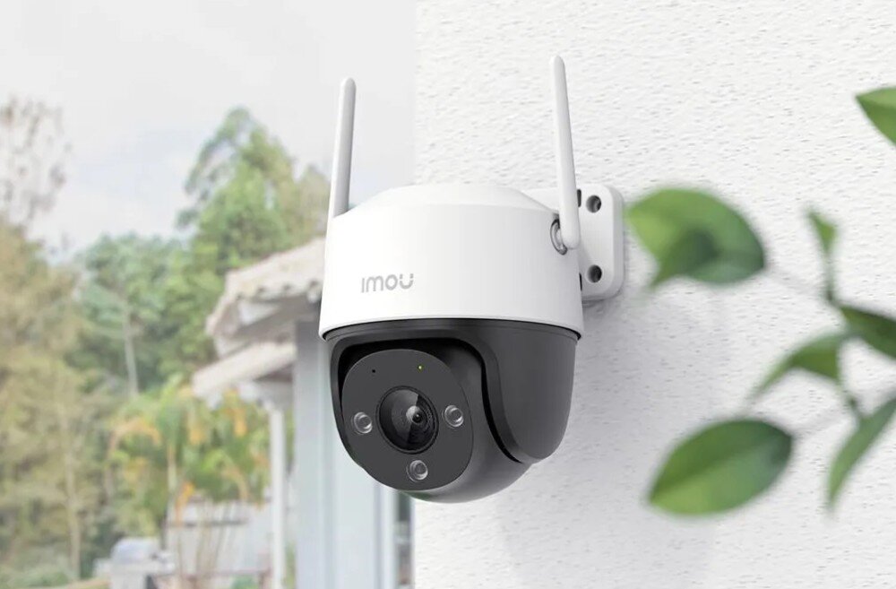 Na obrazie widoczna jest zewnętrzna kamera monitoringu zamontowana na ścianie, z napisem \'IMOU\' na obudowie. W tle znajdują się rozmyte elementy roślinności i budynku.