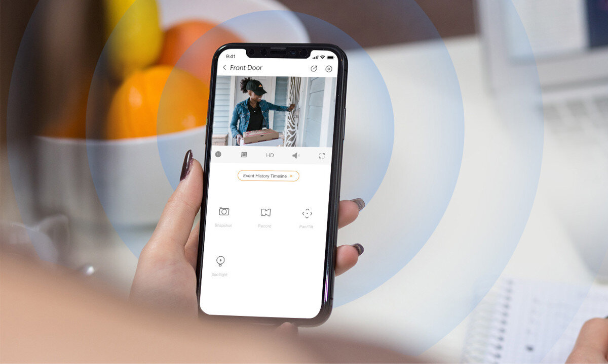 Osoba trzyma smartfon z aplikacją do monitoringu, na ekranie widoczny jest obraz z kamery oraz ikony do zarządzania funkcjami. W tle znajdują się owoce i notatnik na biurku.