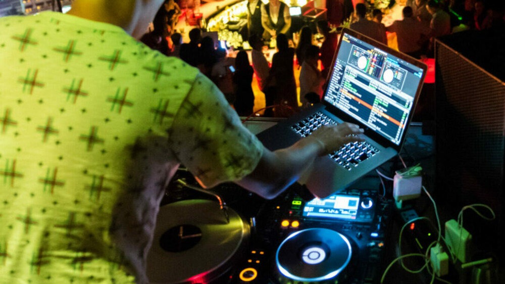 Osoba obsługuje sprzęt DJ-ski, w tym laptop z oprogramowaniem do miksowania muzyki i konsolę DJ-ską. W tle widać rozmytą grupę ludzi na imprezie.