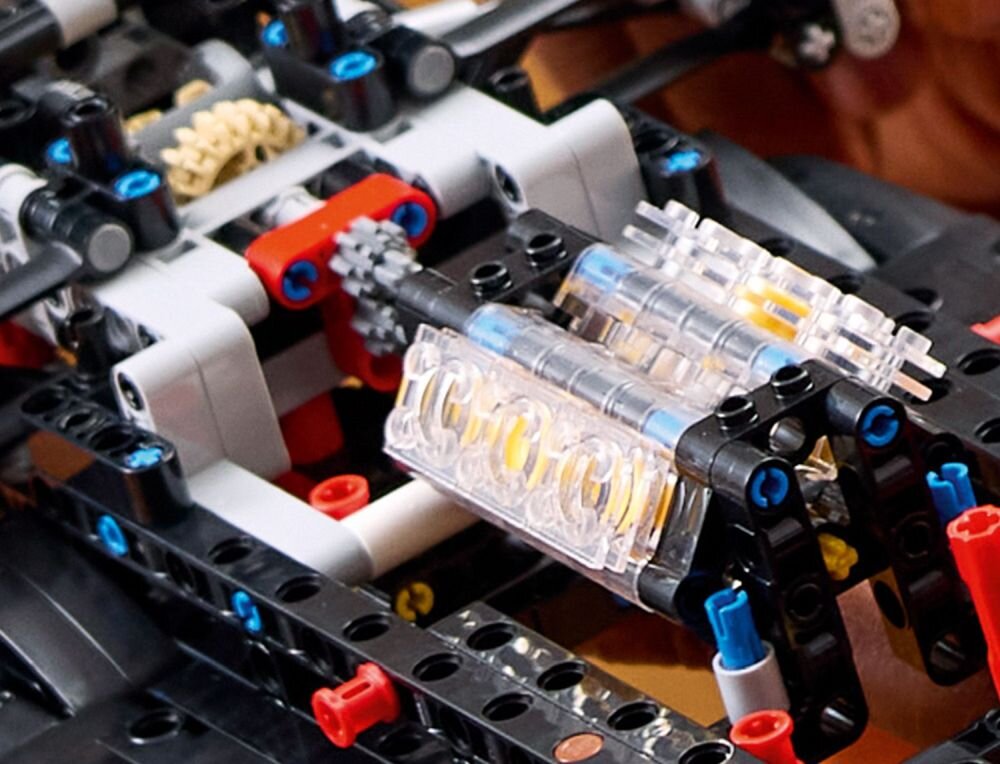 Zbliżenie na model LEGO z serii Technic, przedstawiający skomplikowaną konstrukcję z klocków w różnych kolorach, w tym czarnym, czerwonym i niebieskim. Widoczne są elementy mechaniczne, takie jak zębatki i przezroczyste części.