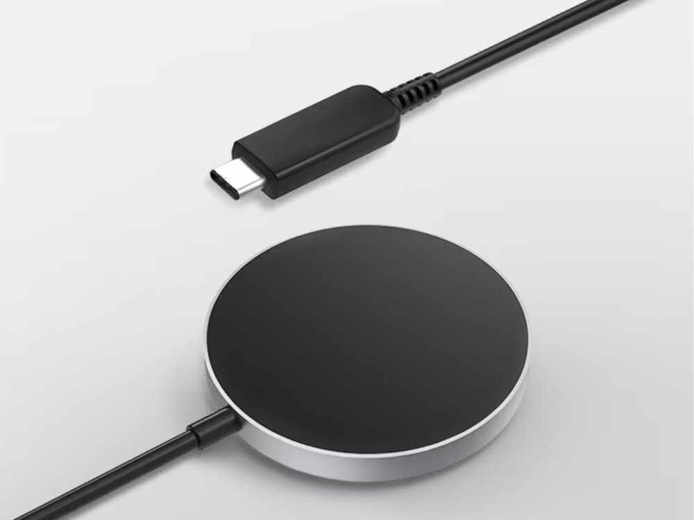 Na obrazie widoczna jest czarna, okrągła ładowarka indukcyjna z kablem zakończonym wtyczką USB-C. Ładowarka ma srebrną obwódkę i leży na jasnym tle.