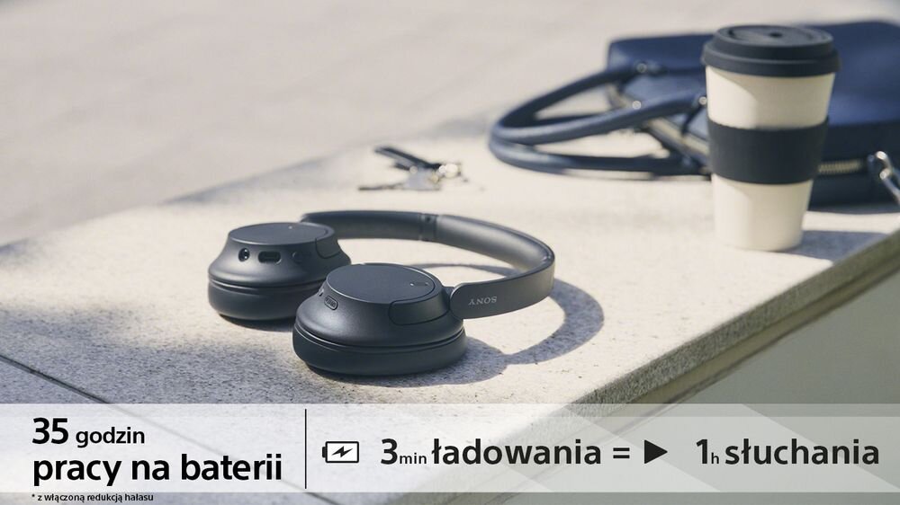Czarne słuchawki nauszne leżą na betonowej powierzchni obok kubka i torby. Tekst na obrazie: \'35 godzin pracy na baterii\', \'3 min ładowania = 1h słuchania\'.