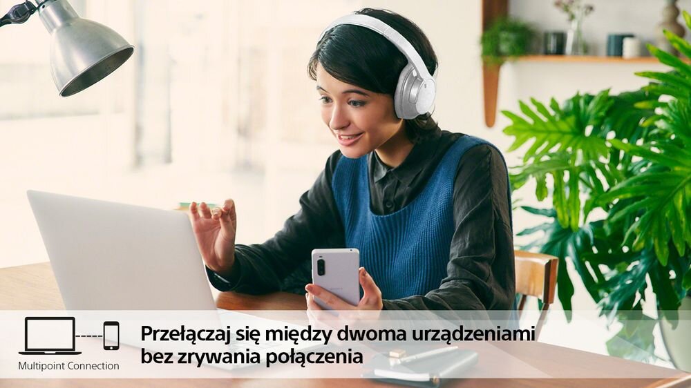 Osoba siedzi przy stole z laptopem i trzyma smartfon, nosząc białe słuchawki nauszne. Na obrazie widoczny jest tekst: \'Przełączaj się między dwoma urządzeniami bez zrywania połączenia\'.