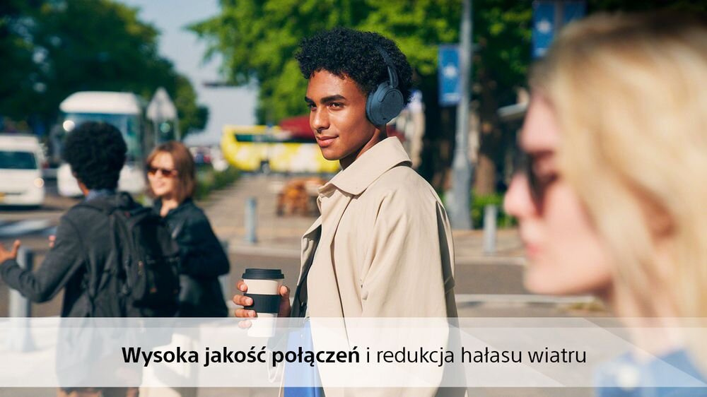 Osoba nosząca czarne słuchawki nauszne stoi na ulicy, trzymając kubek z napojem. Na obrazie widoczny jest tekst: \'Wysoka jakość połączeń i redukcja hałasu wiatru\'.