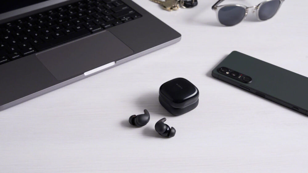Słuchawki dokanałowe SONY LinkBuds WF-LS910 bateria 5,5 h 15,5 h etui szybkie ładowanie 5 minut 1 h, słuchawki z etui ładującym leżące na biurku obok laptopa i telefonu