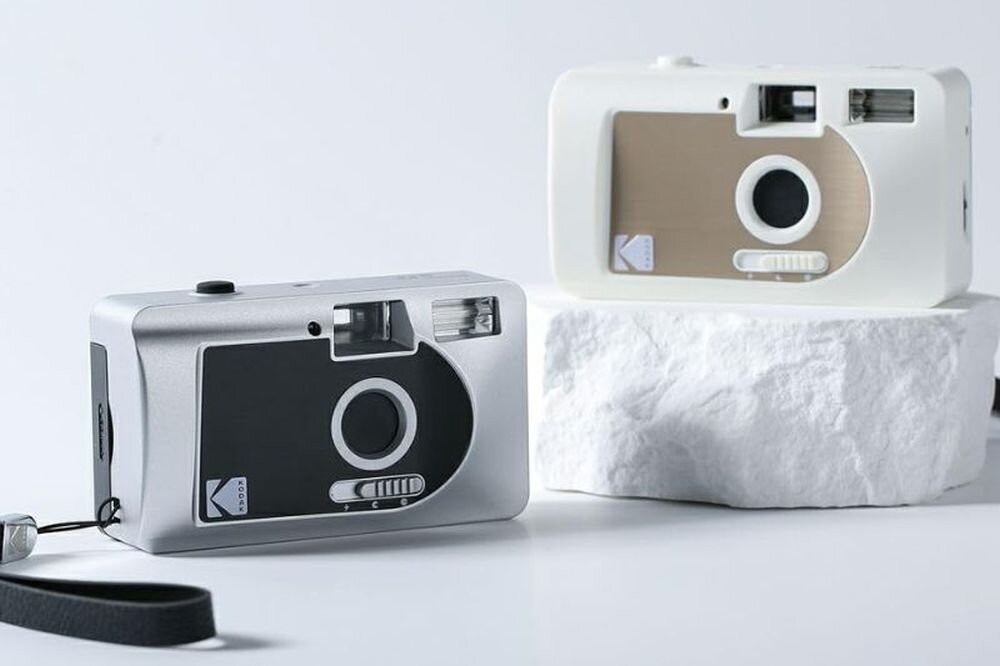 Na obrazie widoczne są dwa aparaty analogowe, jeden srebrny, drugi biały, ustawione na białym tle. Na obu aparatach znajduje się logo \'KODAK\'.