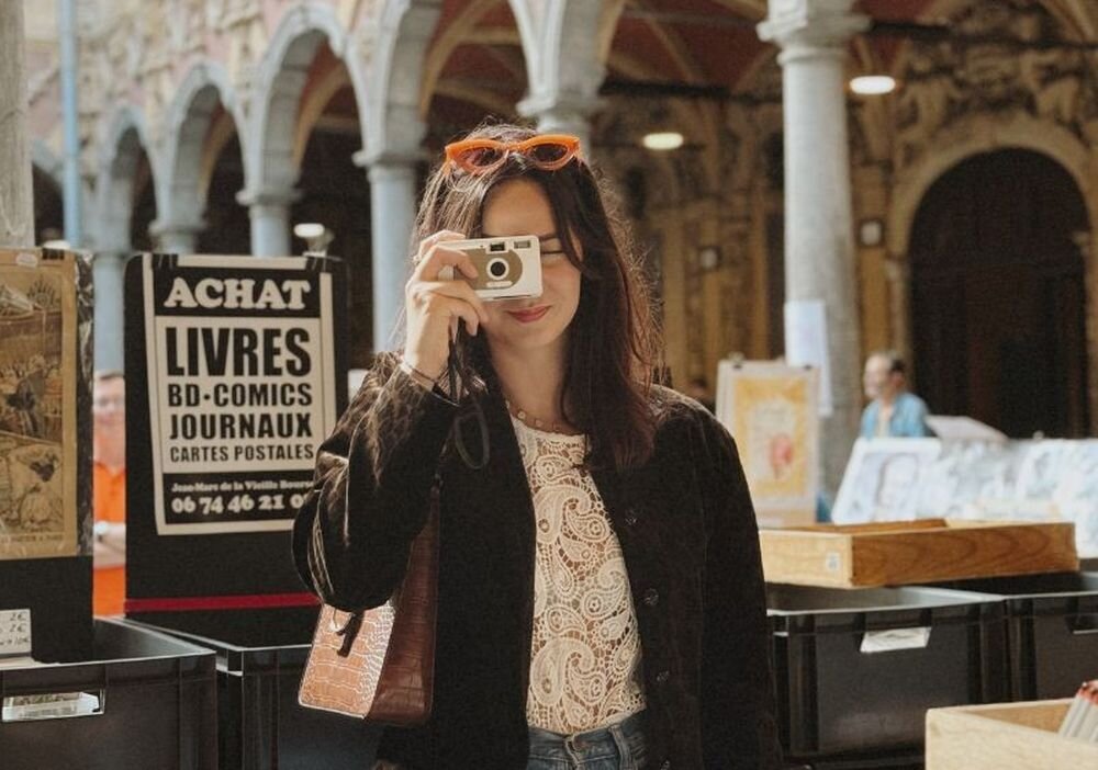 Osoba trzymająca aparat fotograficzny w otoczeniu zadaszonego targu z książkami. Na plakacie widoczny jest tekst: \'ACHAT LIVRES BD-COMICS JOURNAUX CARTES POSTALES\'.