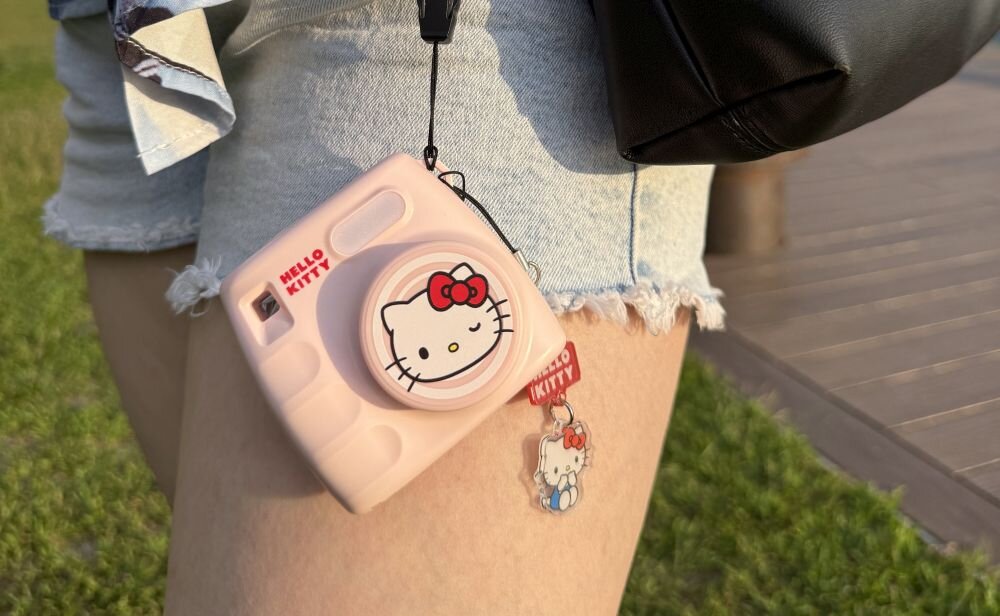 Różowy aparat z motywem Hello Kitty zawieszony na pasku, widoczny na tle jasnych szortów. Na obiektywie znajduje się wizerunek Hello Kitty, a obok wisi breloczek z tym samym motywem.