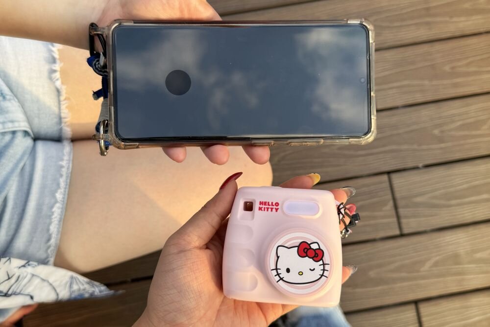 Osoba trzyma różowy aparat z motywem Hello Kitty oraz smartfon w przezroczystym etui. Na aparacie widoczny jest napis \'HELLO KITTY\' oraz rysunek postaci Hello Kitty.