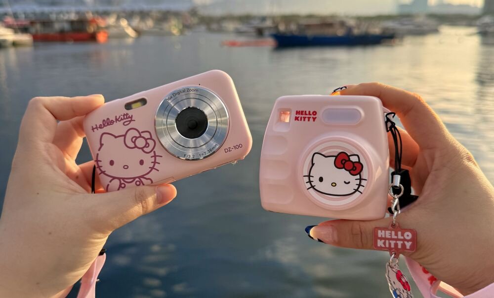 Dwie różowe kamery z motywem Hello Kitty trzymane w dłoniach na tle wody. Na jednej kamerze widoczny jest napis \'Hello Kitty\' oraz rysunek postaci, a na drugiej napis \'Hello Kitty\' i podobny rysunek na obiektywie.