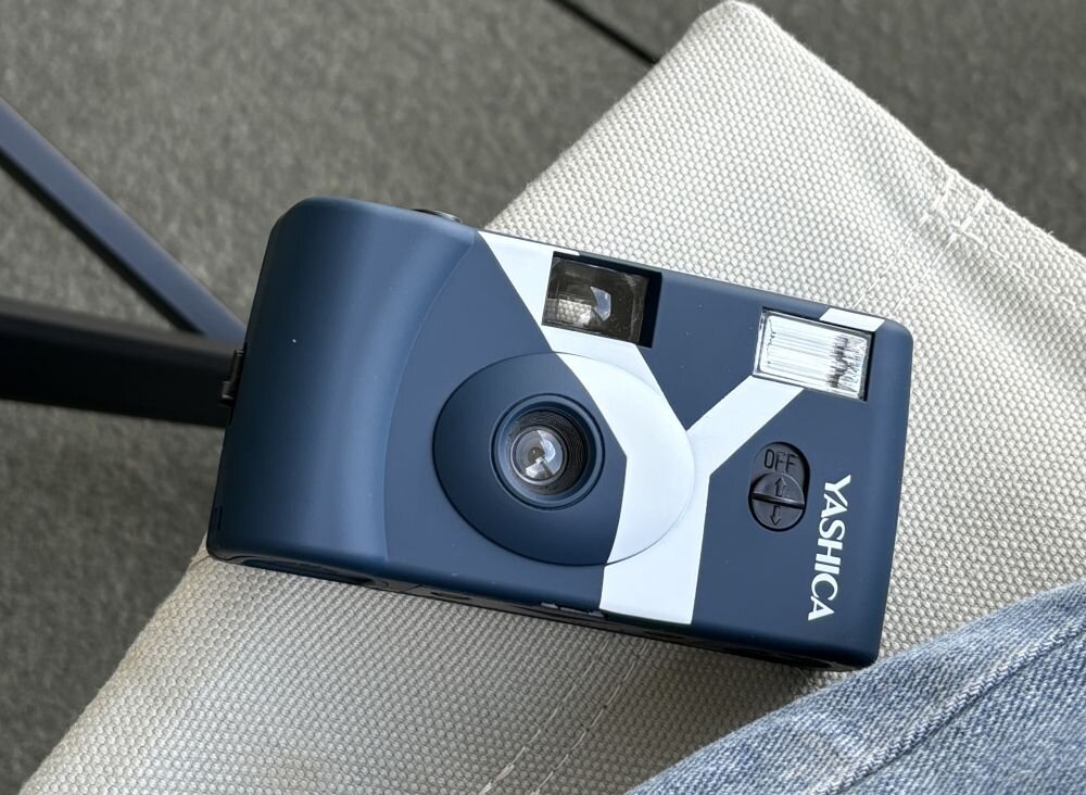Granatowy aparat analogowy z białymi elementami, widoczny obiektyw, lampa błyskowa i napis \'YASHICA\'.