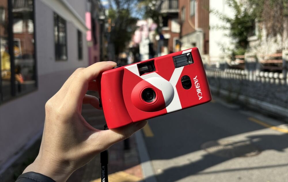 Osoba trzyma czerwony aparat analogowy z białymi elementami i napisem \'YASHICA\'. W tle widoczna jest ulica z budynkami i drzewami.