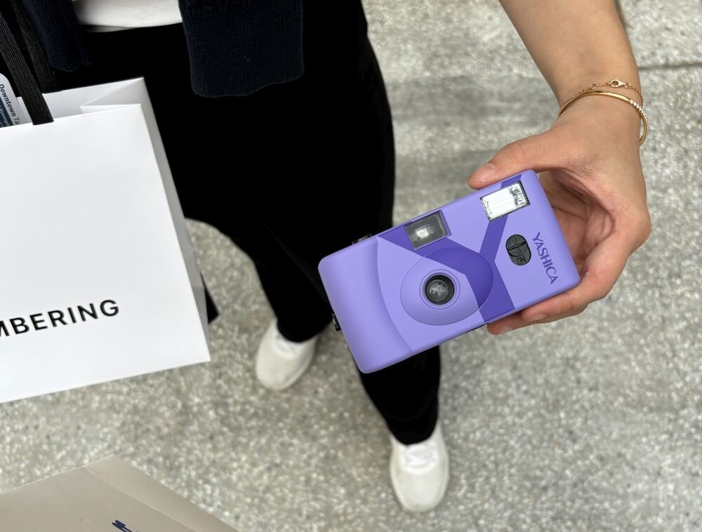 Osoba trzyma fioletowy aparat analogowy z napisem \'YASHICA\'. Obok widoczna jest biała torba z częściowym napisem \'MBERING\'.