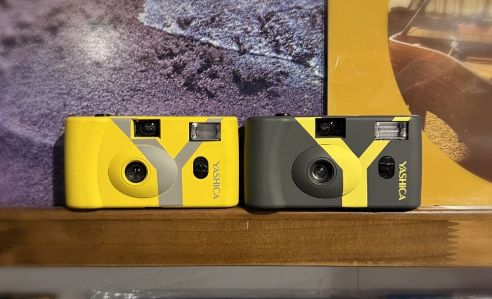 Dwa aparaty analogowe YASHICA w kolorach żółtym i szarym z żółtymi akcentami, umieszczone obok siebie na drewnianej powierzchni. Na obu aparatach widoczne jest logo \'YASHICA\'.