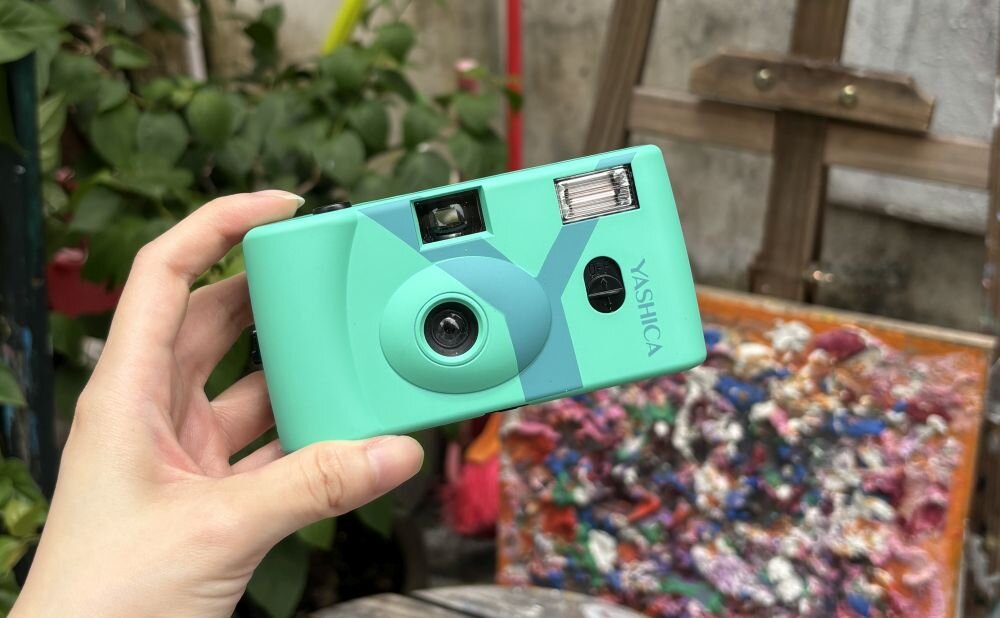 Osoba trzyma aparat analogowy w kolorze granatowym z napisem \'YASHICA\'. W tle widoczne są rośliny i kolorowe płótno na sztaludze.