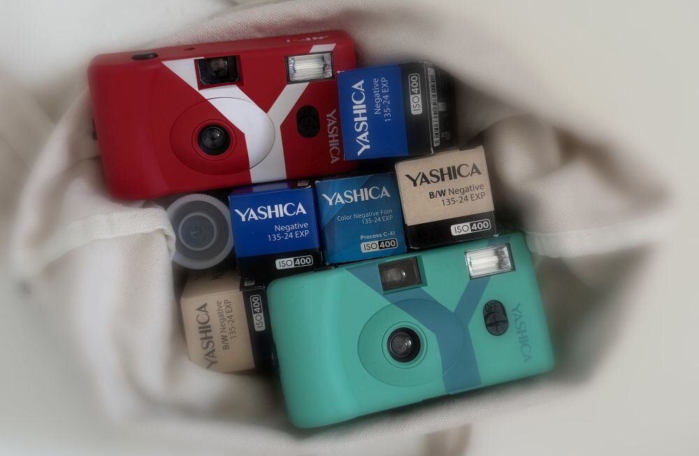 Na obrazie widoczne są dwa aparaty analogowe w kolorach czerwonym i turkusowym oraz kilka pudełek z filmami fotograficznymi marki YASHICA z oznaczeniami ISO 400 i 135-24 EXP.