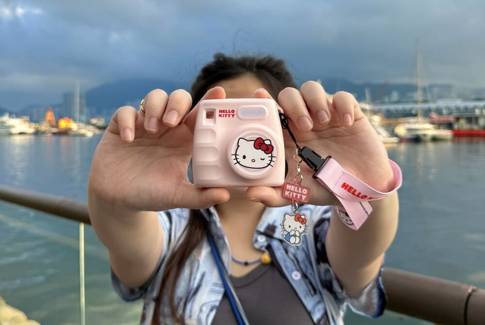 Osoba trzyma różowy aparat z motywem Hello Kitty, z napisem \'HELLO KITTY\' i brelokiem. W tle widać wodę i łodzie.