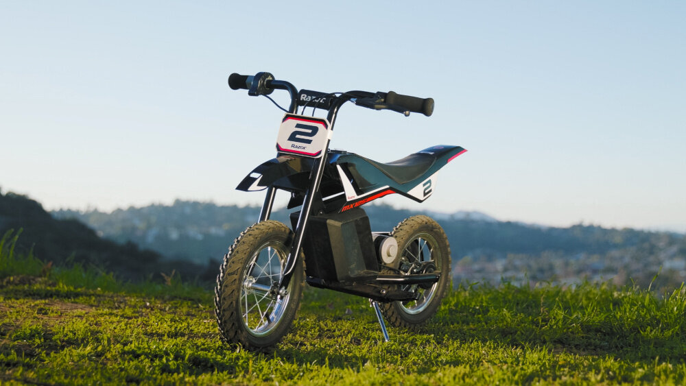 Czarno-czerwony motorek elektryczny z numerem 2 na przodzie, stojący na trawie z widokiem na wzgórza w tle. Na tabliczce widnieje napis \'Razor\'.