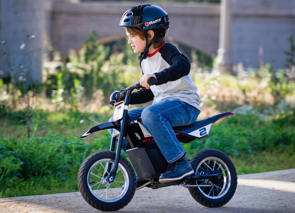 Dziecko jedzie na czarno-czerwonym motorku elektrycznym z numerem 2, nosząc kask z logo \'Razor\'. W tle widoczna jest zieleń i fragment budynku.