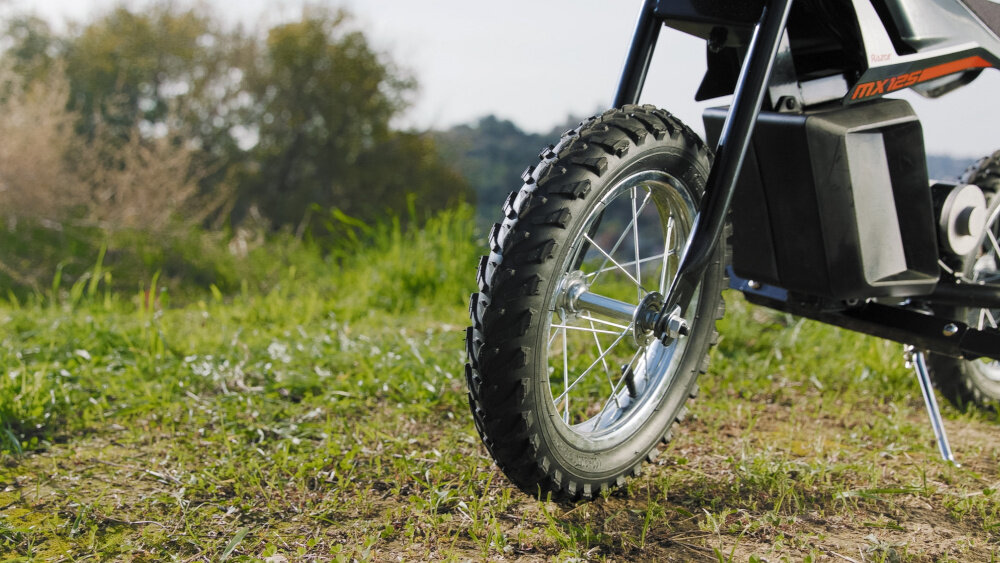 Na obrazie widoczna jest przednia część czarno-czerwonego motorka elektrycznego z grubą oponą terenową na trawiastym podłożu. W tle znajdują się drzewa i krzewy.