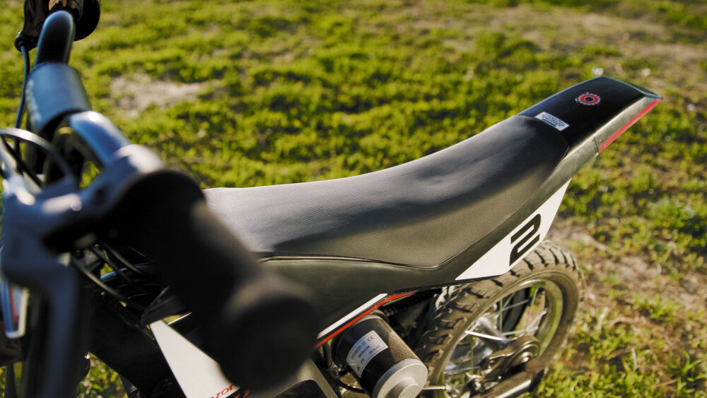 Zdjęcie przedstawia czarno-czerwony motorek elektryczny z widocznym siedzeniem i częścią kierownicy. W tle znajduje się trawiasta powierzchnia.