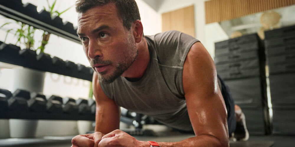 Smartwatch AMAZFIT Balance 2 Czarny Bliskie ujęcie spoconego, brodatego mężczyzny w szarym bezrękawniku, wykonującego ćwiczenie planka (deski) na przedramionach. Patrzy w górę i w prawo. Na jego prawym nadgarstku widać czerwony pasek zegarka. W tle regały z hantlami i stosy mat