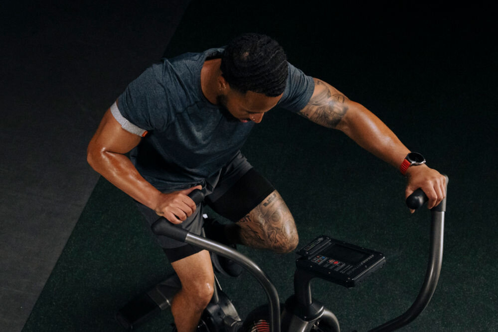 Smartwatch AMAZFIT Balance 2 Czarny Zdjęcie z góry przedstawiające spoconego, umięśnionego mężczyznę w szarej koszulce i czarnych spodenkach, jadącego na rowerze treningowym typu air bike w słabo oświetlonej siłowni. Mężczyzna ma tatuaże na ręce i udzie oraz czerwony zegarek sportowy na lewym nadgarstku