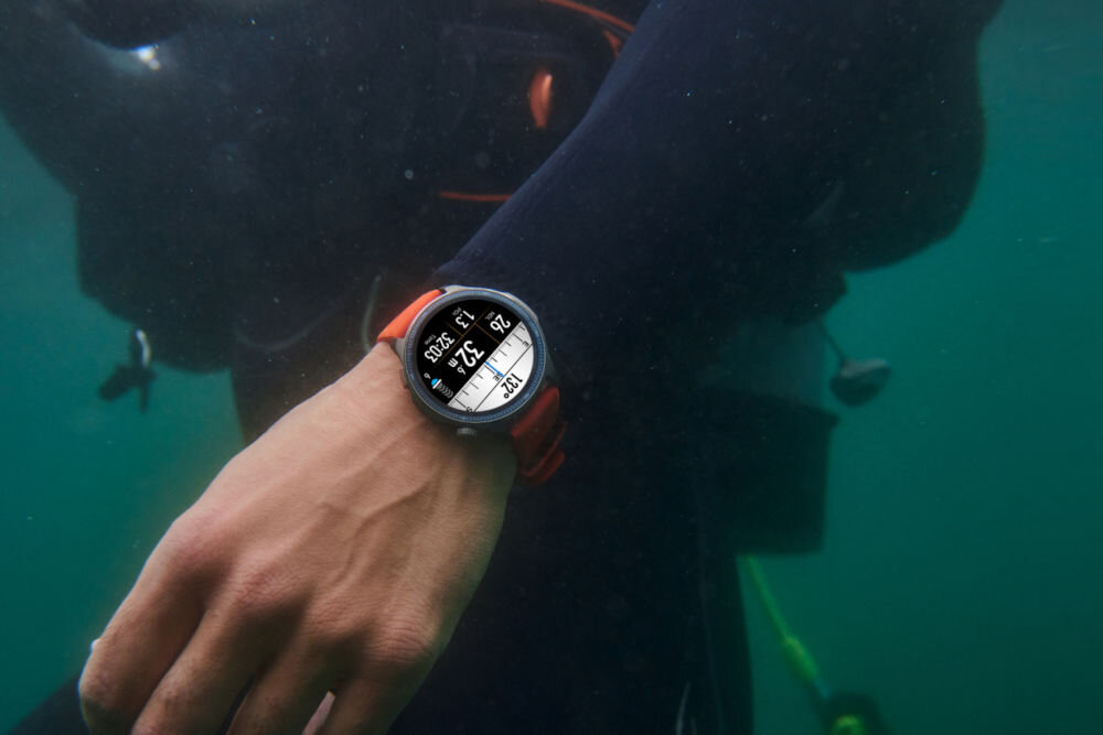 Smartwatch AMAZFIT Balance 2 Czarny Zbliżenie na rękę w czarnym kombinezonie do nurkowania z okrągłym zegarkiem sportowym z pomarańczową opaską, zanurzoną w zielonkawej wodzie. Na ekranie zegarka widoczne są dane, takie jak głębokość (32 metry) i czas