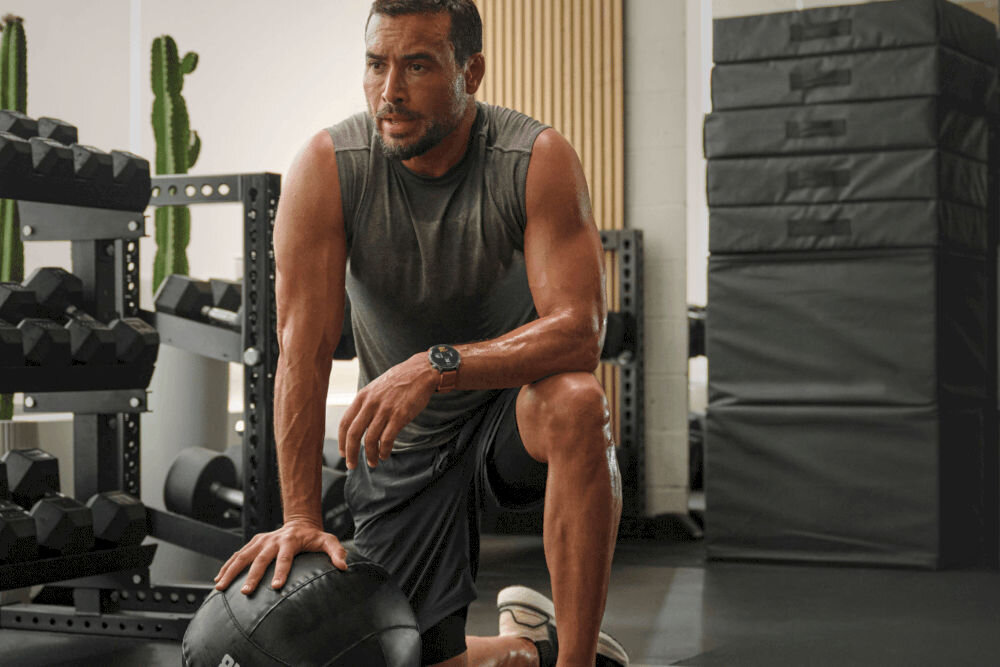 Smartwatch AMAZFIT Balance 2 Czarny Mężczyzna klęczący na jednym kolanie na podłodze siłowni, opierający się o czarną piłkę lekarską. Jest spocony i ma na sobie szary bezrękawnik i czarne szorty. Na jego lewym nadgarstku widoczny jest okrągły zegarek w ciemnym kolorze. W tle regały z ciężarami i ułożone w stosy czarne skrzynki