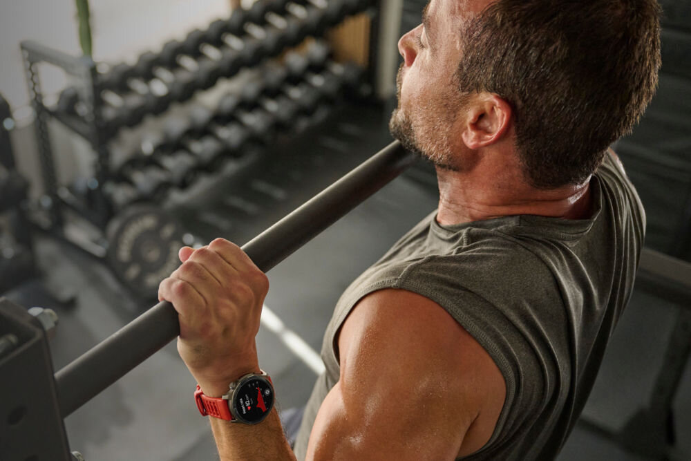 Smartwatch AMAZFIT Balance 2 Czarny Zbliżenie z boku na spoconego, brodatego mężczyznę w szarym bezrękawniku, który trzyma oburącz metalowy drążek. Na jego prawym nadgarstku widać duży, okrągły zegarek sportowy z czerwoną opaską. Tło to zamazane wnętrze siłowni.