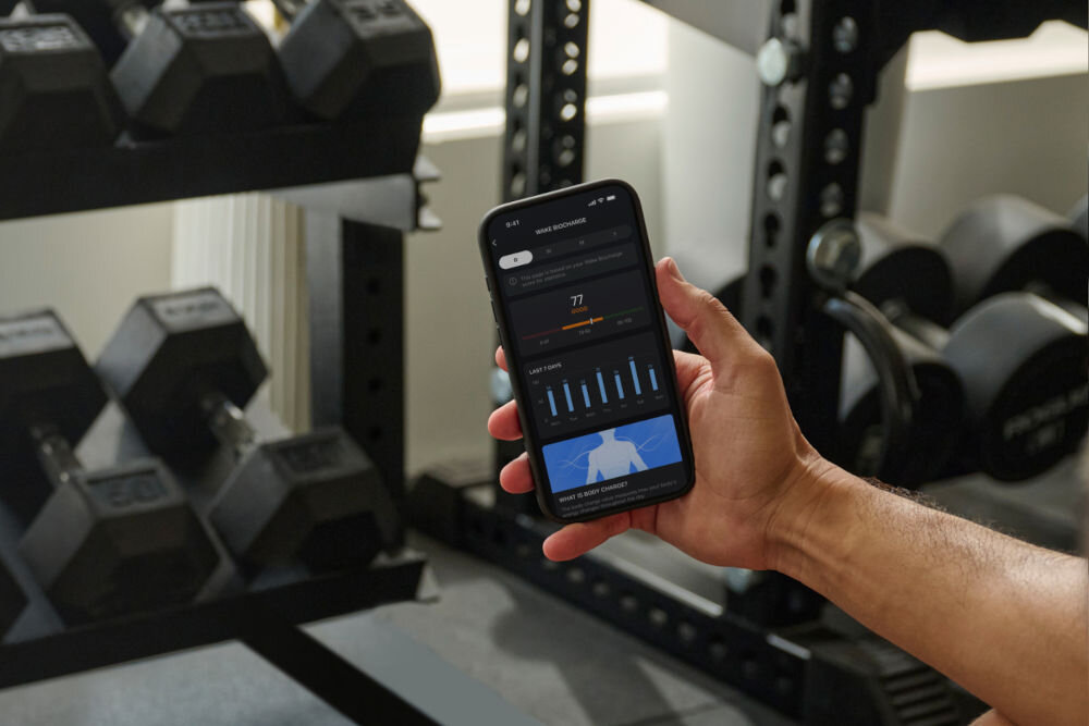 Smartwatch AMAZFIT Balance 2 Czarny Zbielone ujęcie dłoni trzymającej smartfona, na którego ekranie widać aplikację fitness wyświetlającą dane dotyczące regeneracji (Wake Biocharge) z wynikiem 77 i wykresami słupkowymi. Tło to siłownia z metalowym regałem i rzędami hantli