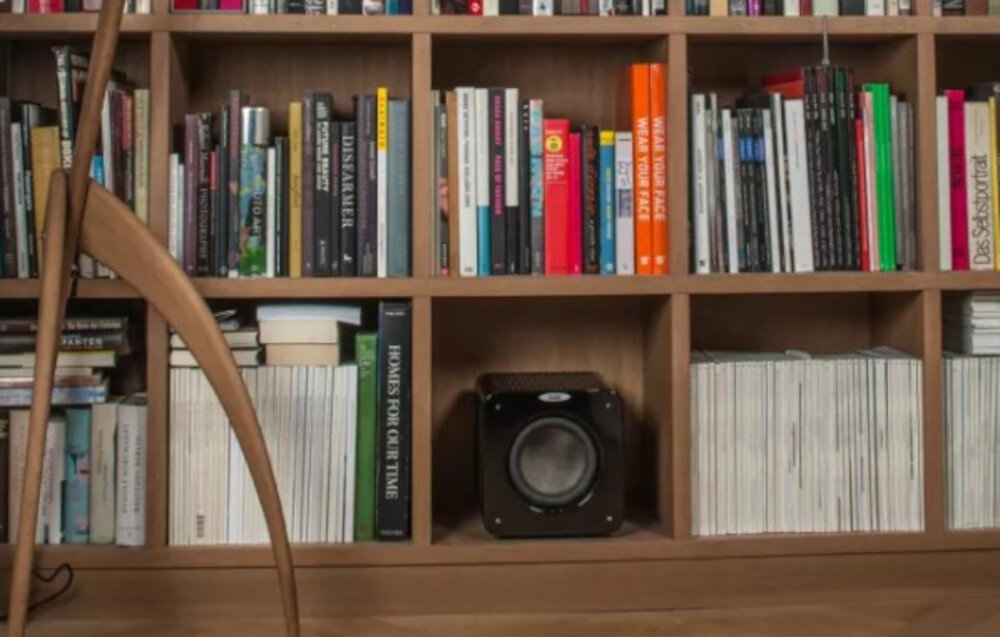 Subwoofer VELODYNE MicroVee X 6.5 design kompaktowy, solidna konstrukcja, subwoofer stojący, Czarny kompaktowy subwoofer VELODYNE MicroVee X 6.5 ustawiony na półce wśród książek i magazynów, w domowym salonie, pokazujący integrację sprzętu audio z wnętrzem
