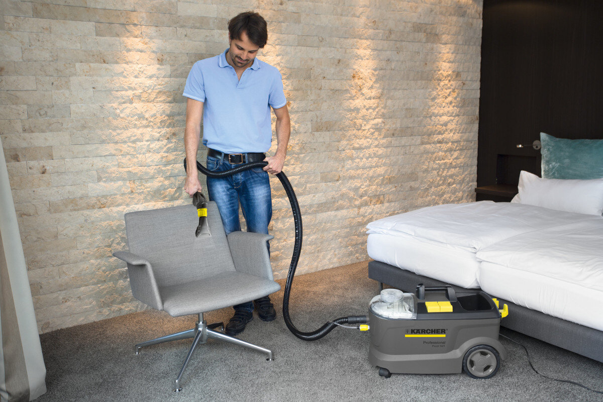 Mężczyzna używa urządzenia ekstrakcyjnego do czyszczenia szarego fotela w pokoju z łóżkiem. Na urządzeniu widoczne jest logo \'KARCHER\'.