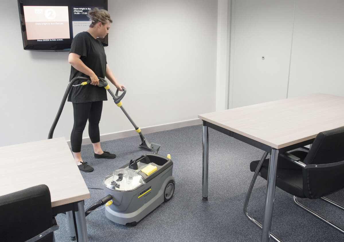 Osoba używa urządzenia ekstrakcyjnego KARCHER na wykładzinie w pomieszczeniu biurowym. W tle widoczne są dwa stoły i krzesła oraz ekran na ścianie.