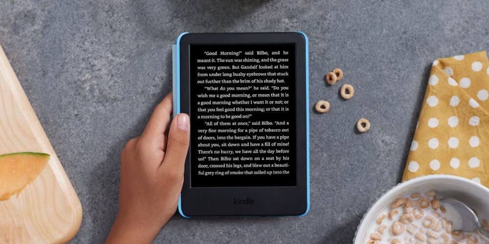 Czytnik e-booków AMAZON Kindle Kids Unicorn Valley 6 cali, Podświetlany ekran, Wi-Fi, Bez reklam Kindle Kids z wyświetlonym tekstem książki leży na blacie obok miski z płatkami, ściereczki w kropki i deski z kawałkiem melona waga kolorowa okładka ochrona obudowy indywidualny charakter