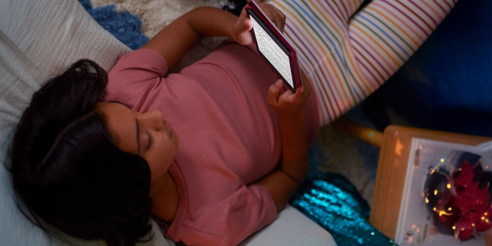 Czytnik e-booków AMAZON Kindle Kids Space Whale 6 cali, Podświetlany ekran, Wi-Fi, Bez reklam Dziewczynka leży wieczorem na podłodze lub łóżku i czyta rozświetlony ekran Kindle Kids w różowym etui, w tle kolorowe elementy wystroju pokoju gniazdo USB-C ładowanie baterii przesyłanie książek jedno pełne ładowanie wystarcza na wiele tygodni ogranicza potrzebę częstego ładowania praktyczny kompan na wyjazdy