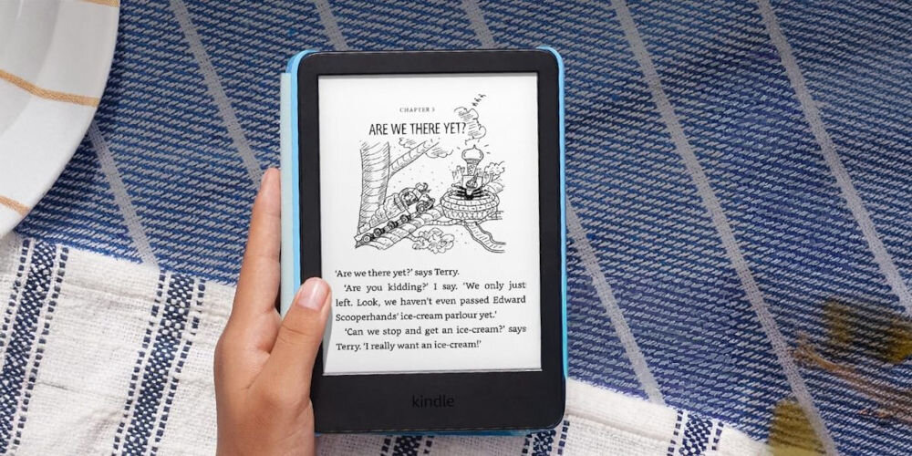 Czytnik e-booków AMAZON Kindle Kids Space Whale 6 cali, Podświetlany ekran, Wi-Fi, Bez reklam Kindle Kids z czarno-białą ilustracją i tekstem trzymany w dłoni nad niebiesko-białym, pasiastym kocem piknikowym wyświetlacz E-Ink ograniczenie emisji światła i poboru energii wyraźny tekst sprzyja skupieniu