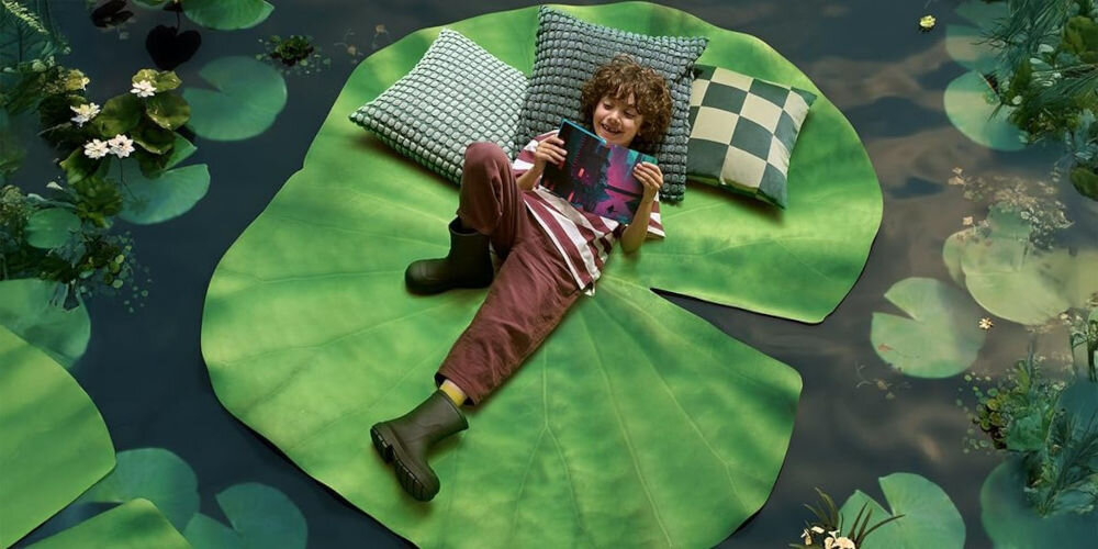 Czytnik e-booków AMAZON Kindle Paperwhite Kids Diary of a Wimpy Kid 7 cali, Podświetlany ekran, Wi-Fi, Bez reklam dziecko leży na ogromnym liściu otoczonym poduszkami i trzyma kolorowe etui Kindle 16 GB pamięci synchronizacja z chmurą Amazon Wi-Fi Bluetooth audiobooki subskrypcja Amazon Kids+ zawartość filtrowana wiekowo