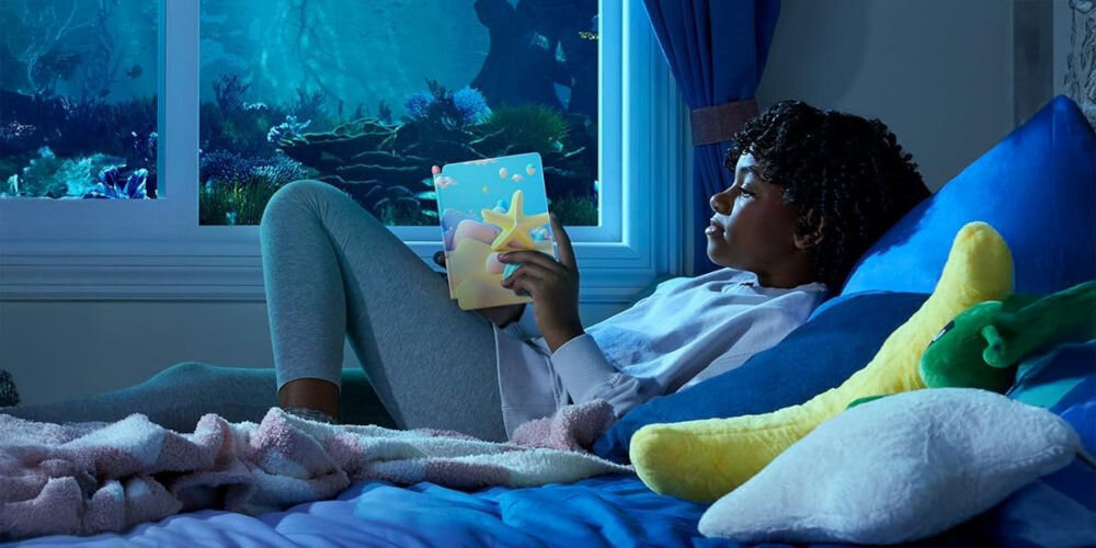 Czytnik e-booków AMAZON Kindle Paperwhite Kids Diary of a Wimpy Kid 7 cali, Podświetlany ekran, Wi-Fi, Bez reklam Dziewczynka leży wieczorem w łóżku, trzyma Kindle Paperwhite Kids z etui Starfish, w tle okno z podwodnym motywem kontrola nad treściami Parent Dashboard brak reklam narzędzia edukacyjne wsparcie rozwoju zdrowe nawyki czytelnicze